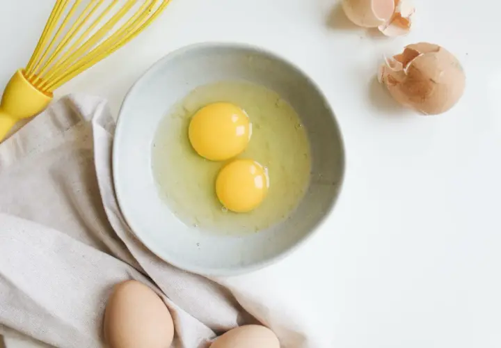 Deux jaunes d'œufs dans un bol, prêts à être battus. Une question se pose : peut-on congeler des jaunes d'œufs ? Un fouet jaune et des coquilles d'œufs complètent la scène.