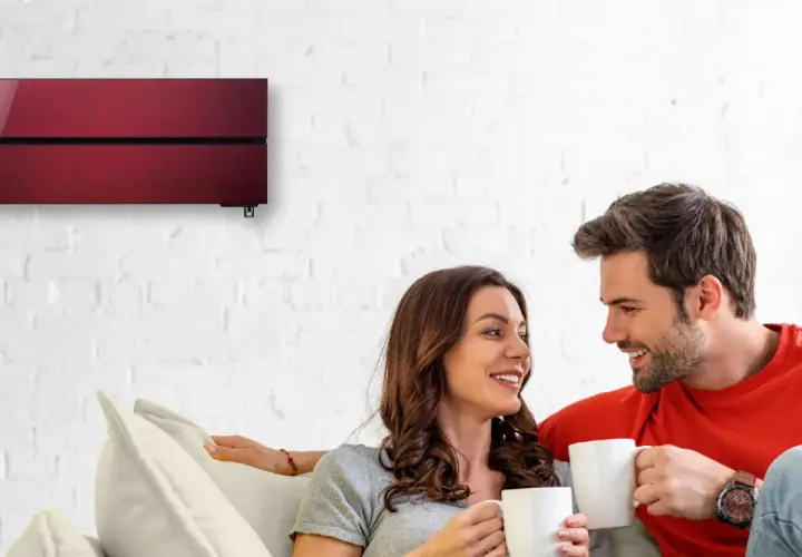 Pareja sonriente en un sofá, disfrutando de café. Arriba, un aire acondicionado rojo moderno garantiza cop eficiencia.
