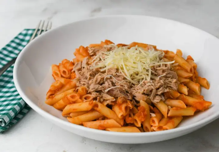 Macarrones con tomate y atún desmenuzado, cubiertos con queso rallado, servidos en un plato blanco.