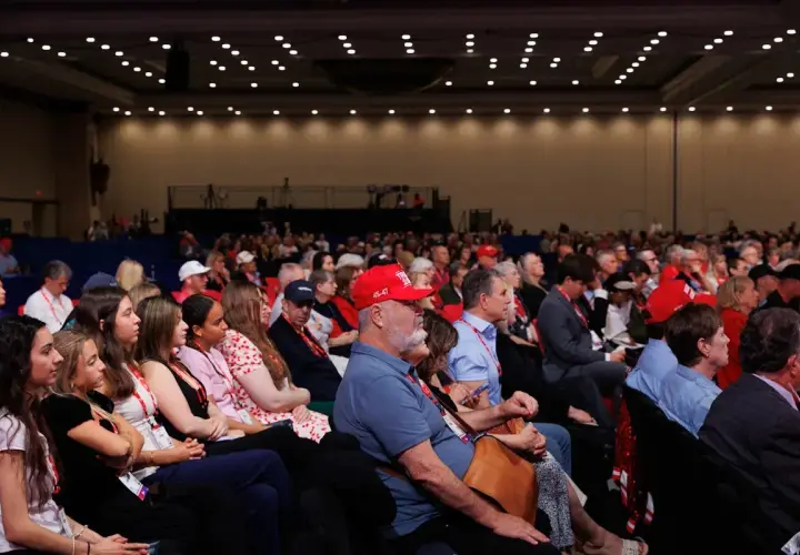 A large audience attends a government conference, with many people wearing red hats.