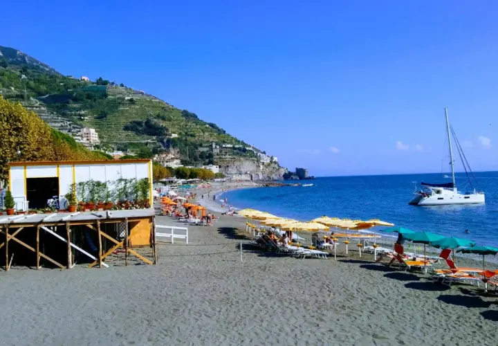 Plage de sable noir à Maiori, Italie, avec parasols, chaises longues, un catamaran blanc et des collines verdoyantes.