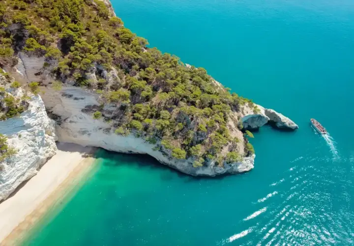 Widok z lotu ptaka na skaliste wybrzeże z piaszczystą plażą i turkusową wodą. Statek wycieczkowy płynie w stronę łuku skalnego. Idealne miejsce, gdzie do Włoch!