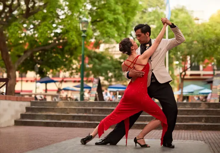 Coppia di ballerini latino americani in un appassionato tango all'aperto, lei in abito rosso, lui in giacca chiara.