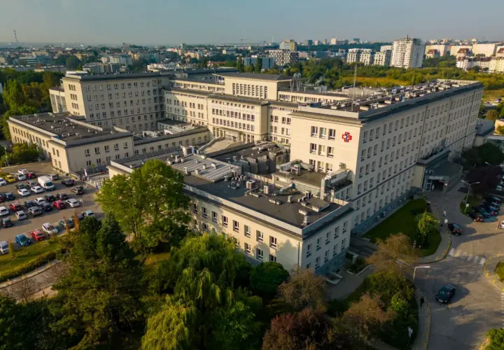 Widok z lotu ptaka na duży kompleks szpitalny z czerwonym krzyżem na fasadzie, otoczony drzewami i parkingiem.