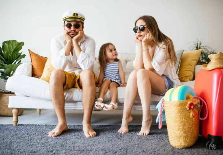 Famille dépitée sur un canapé, valise rouge prête. Annulation location vacances cas de force majeure, les vacances rêvées s'envolent.