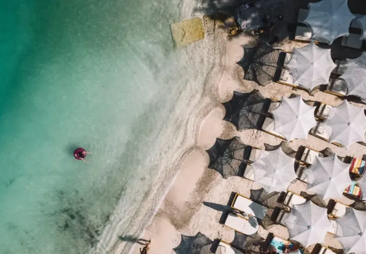 Widok z lotu ptaka na turkusowe morze i piaszczyste plaże Albanii. Jedna osoba relaksuje się na dmuchanym kole, a obok rzędy białych parasoli i leżaków.