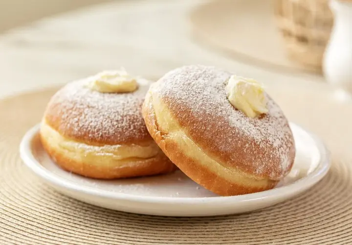 Due bomboloni ripieni di crema, spolverati di zucchero a velo, mostrano la differenza tra bombolone e krapfen: il primo è più soffice e senza buco.