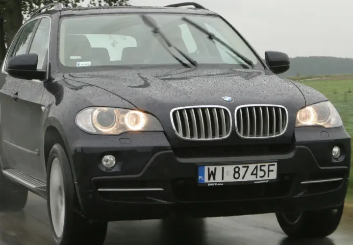 Ciemny BMW E70 na mokrej drodze, w tle zielone pola i drzewa.