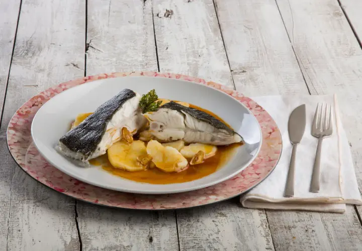Rodaballo gallego servido con patatas y salsa, acompañado de cubiertos y servilleta sobre una mesa de madera.