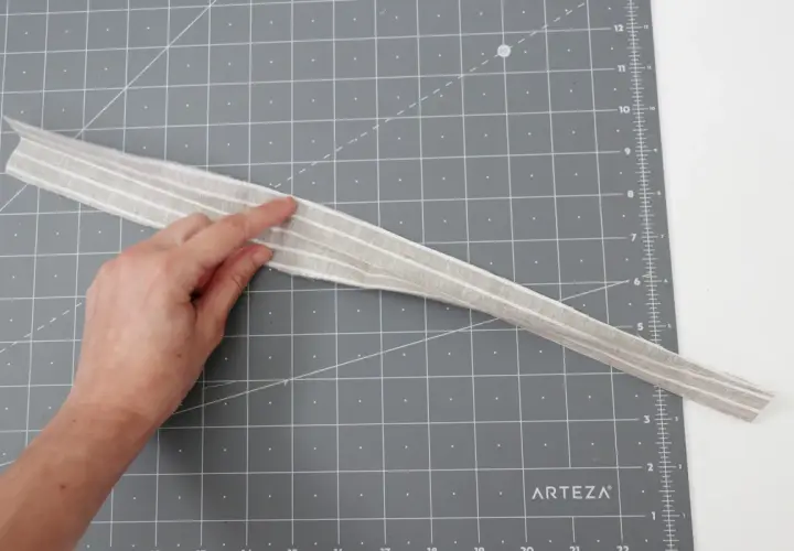 A hand points to a striped fabric strip, possibly for a belt loop pattern, on a cutting mat.