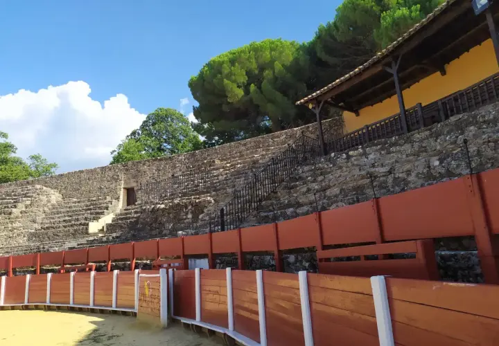 La plaza de toros cuadrada en España, con sus gradas de piedra y barreras de madera, espera la emoción bajo un cielo azul y nubes algodonosas.