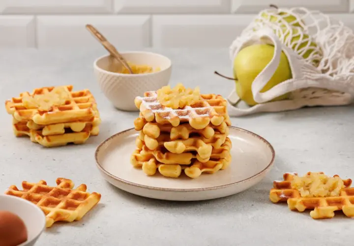 Goldbraune Waffeln mit Apfelmus bestreut, daneben eine Schüssel mit Apfelmus und ein grüner Apfel im Netzbeutel.