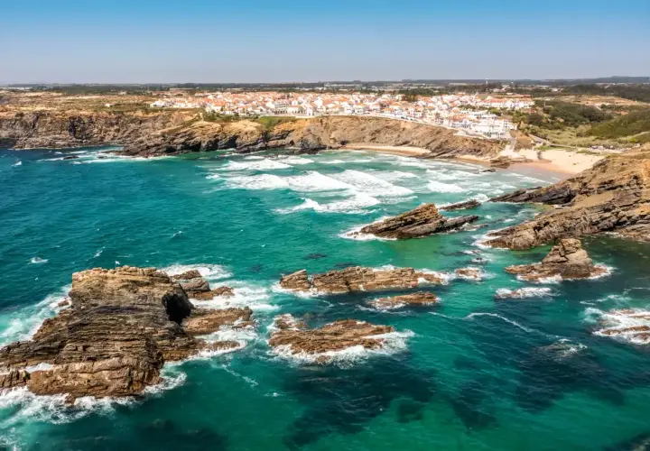 Costa rocosa y aguas turquesas en una **playa Portugal**. Un pueblo blanco se asoma al acantilado.