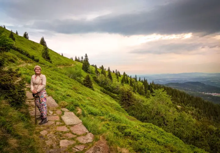 Kobieta z kijkami na kamiennym szlaku, podziwia widok na zielone zbocze i las. Widać, ile się wchodzi na Śnieżkę.