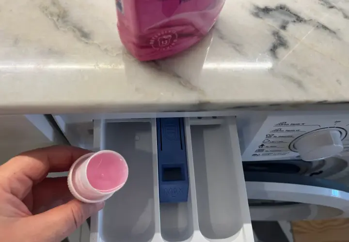 Hand hält Dosierkappe mit rosa Flüssigkeit. Waschpulver direkt in die Trommel geben? Nein, hier in die Schublade.