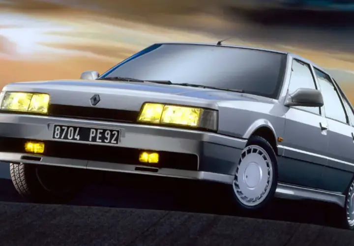 Un elegante Renault 21 Turbo plateado, con sus faros amarillos encendidos, se luce contra un cielo crepuscular.