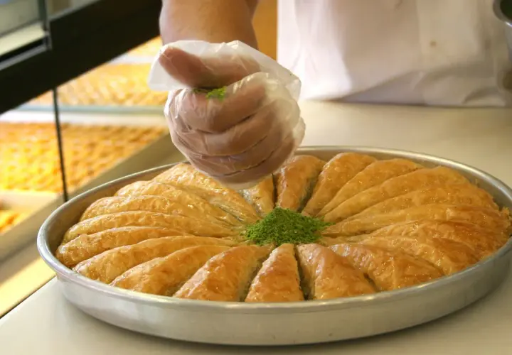 Ręka w rękawiczce posypuje zielonymi pistacjami okrągłą blachę pełną złocistej baklawy. To właśnie baklava co to za deser!