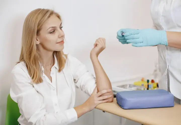 Kobieta z blond włosami w białej koszuli i pielęgniarka w rękawiczkach przygotowują się do pobrania krwi.