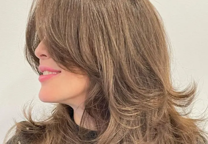 Mujer sonríe con un corte mariposa en su cabello castaño, con capas que enmarcan su rostro.