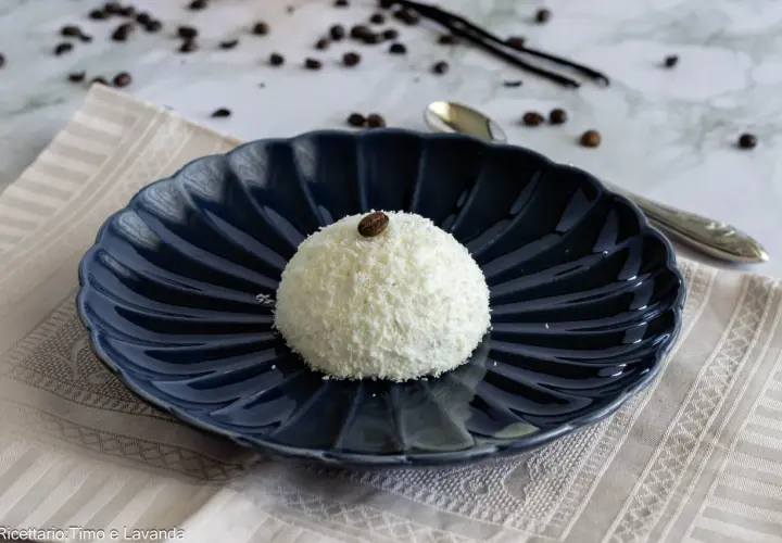 Semifreddo tartufo bianco, un dolce raffinato ricoperto di cocco e decorato con un chicco di caffè, su un piatto blu scuro.