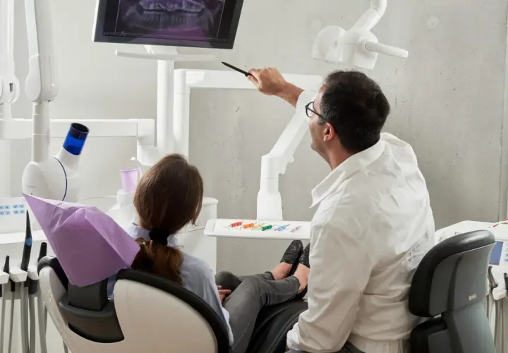 Dentysta wskazuje na ekran z prześwietleniem zębów pacjentce siedzącej na fotelu.