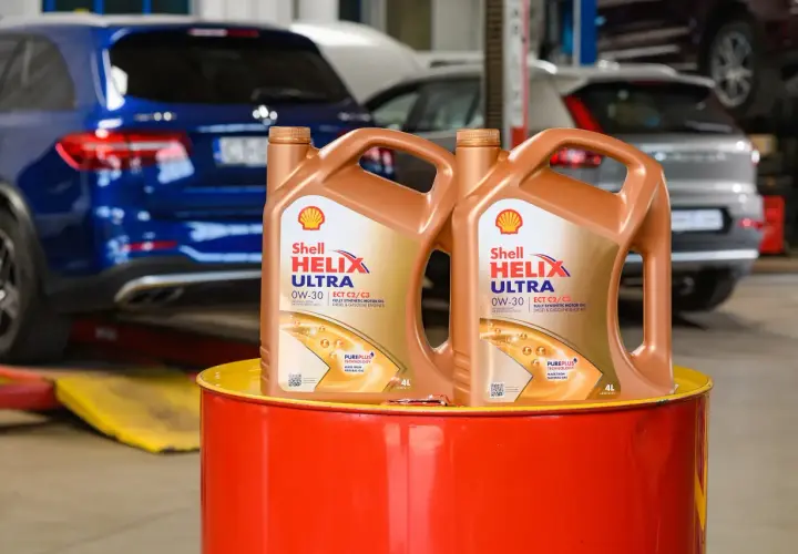 Dwa kanistry oleju silnikowego Shell Helix Ultra 0W-30 stoją na czerwonym bębnie. W tle widać samochody w warsztacie.
