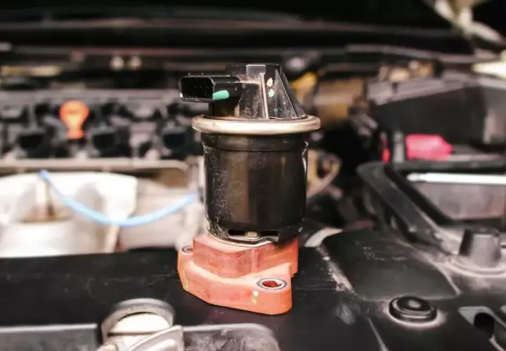 Componente negro con conector eléctrico, posiblemente una válvula de admisión, sobre una base rojiza en un motor.