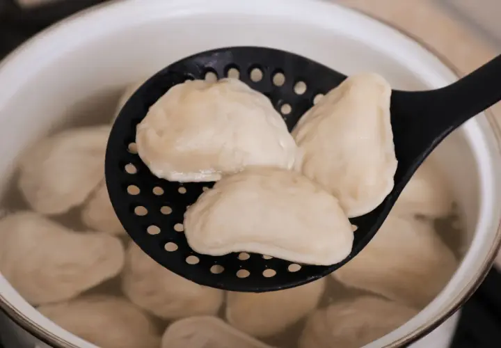 Gotowane pierogi na czarnej łyżce cedzakowej w garnku z wodą.
