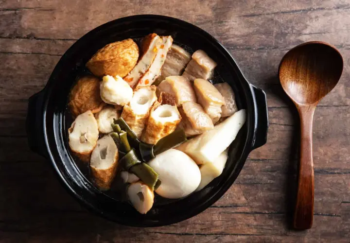 A steaming pot of oden, a Japanese fish cake stew, with various ingredients like daikon and pork belly, served with a wooden spoon.