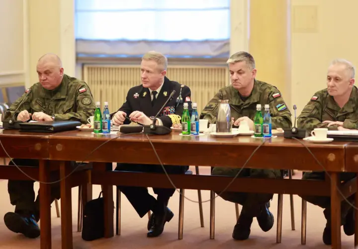 Czterech mężczyzn w mundurach siedzi przy drewnianym stole, przed nimi butelki wody i filiżanki.