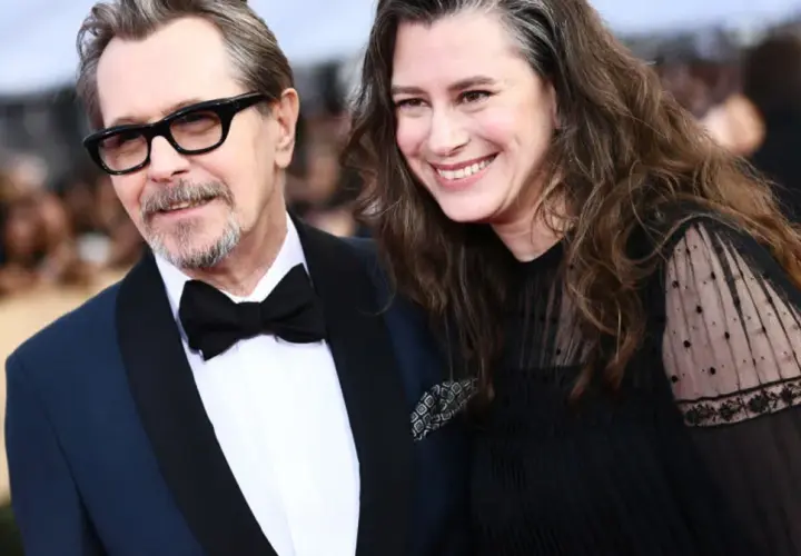 Gary Oldman z żoną, oboje uśmiechnięci, on w eleganckim garniturze, ona w czarnej sukni z koronkowymi rękawami.