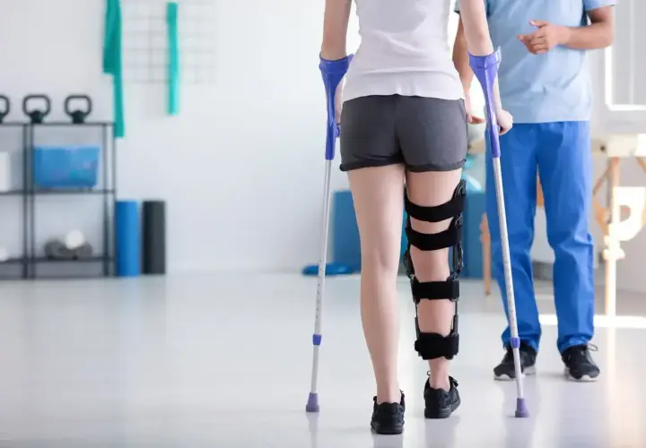 Pacjentka z ortezą na nodze uczy się chodzić o kulach pod okiem fizjoterapeuty w gabinecie rehabilitacyjnym.