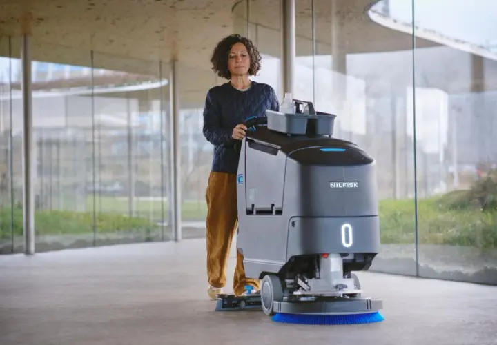 Mujer operando una máquina de Nilfisk, parte de sus avanzados sistemas de limpieza, en un pasillo moderno con grandes ventanales.