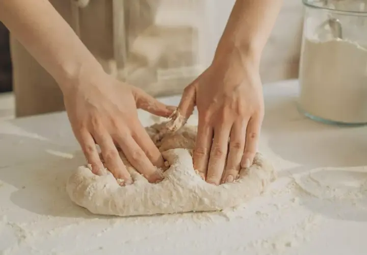 Manos amasando masa pan sobre una mesa con harina esparcida. Un frasco de vidrio con harina y un cuenco están al fondo.
