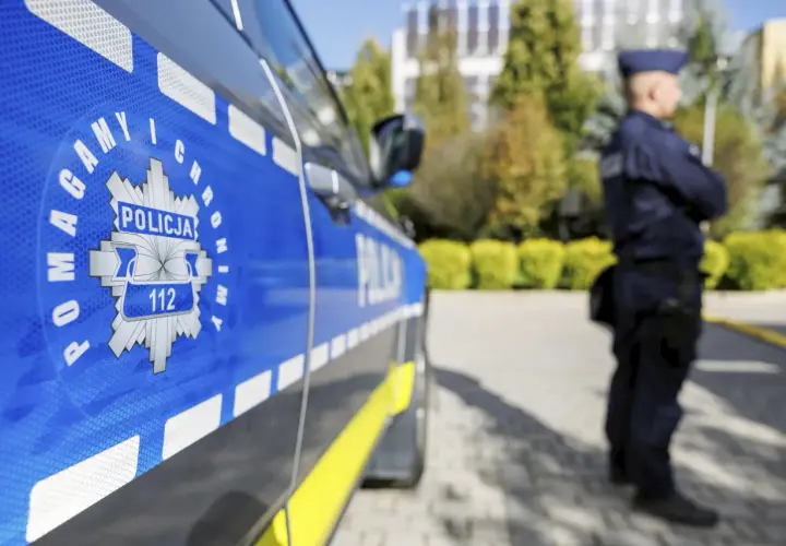Policjant w mundurze stoi obok radiowozu z napisem "POLICJA" i numerem alarmowym 112.