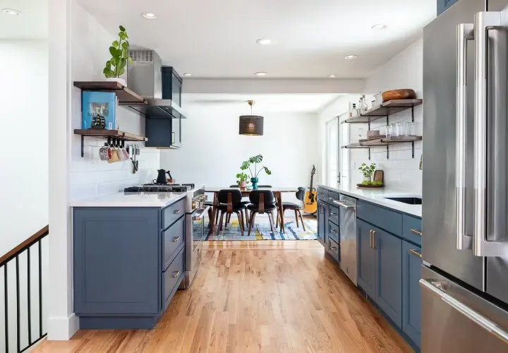 A modern kitchen with deep blue cabinets, white subway tile backsplash, and warm wood floors. The cabinet colors offer a stylish contrast to the stainless steel appliances.