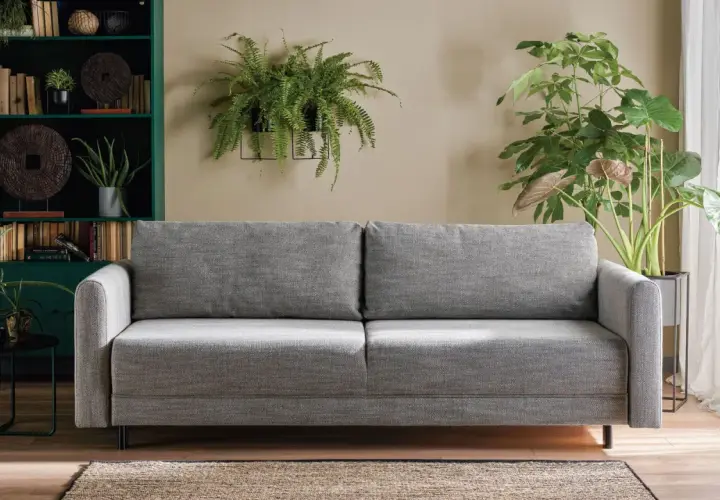 Sofa w kolorze popielaty jaki to kolor, otoczona zieloną biblioteczką i bujną roślinnością.