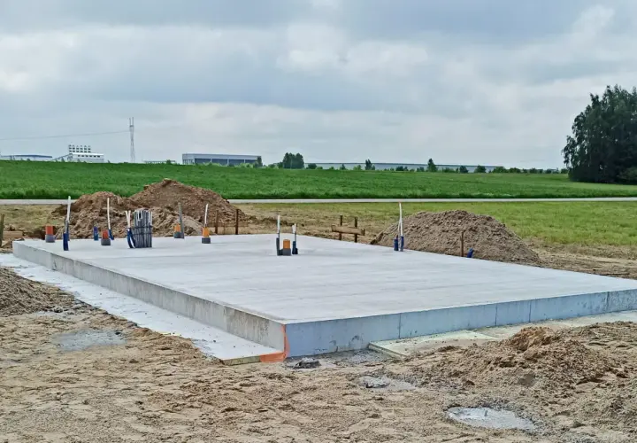 Gotowy fundament pod dom szkieletowy, z wystającymi rurami i zbrojeniem, otoczony piaskiem i zielonym polem.