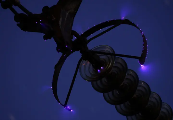 Des arcs électriques violets, les feux de Saint-Elme, crépitent sur une structure métallique et des isolateurs en verre contre un ciel nocturne.