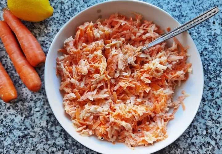 Pyszna surówka z marchewki jak z restauracji, gotowa do podania. Obok leżą świeże marchewki i cytryna.