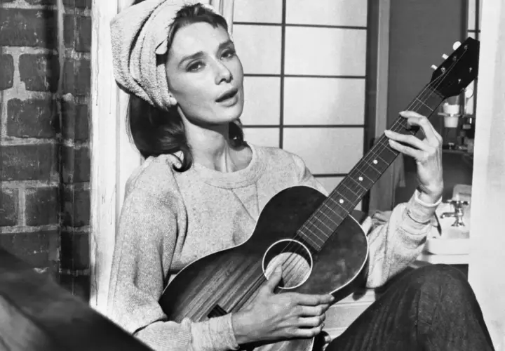 Audrey Hepburn, z ręcznikiem na głowie, gra na gitarze, śpiewając "Moon River".