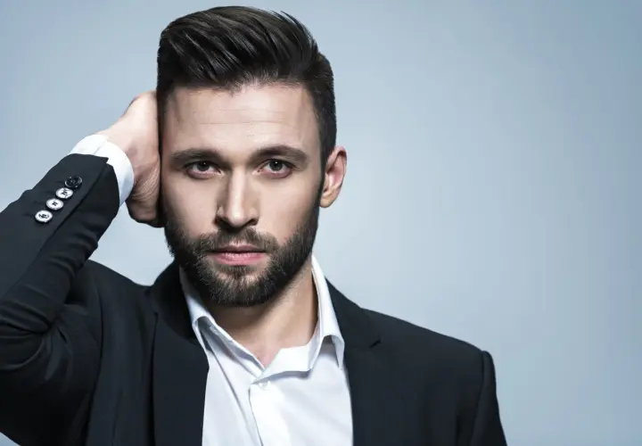Hombre con un peinado con tupe, barba y traje negro, tocándose la cabeza.
