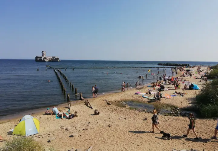 Słoneczna babie doły plaża tętni życiem. Ludzie opalają się, kąpią w morzu, a w oddali widać zabytkowy fort.