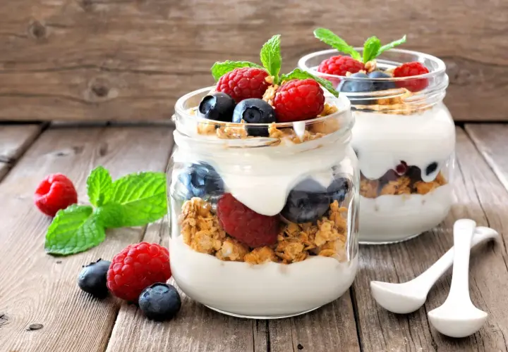 Dos frascos de vidrio con yogur, granola, frambuesas y arándanos. ¡Cuidado! El yogur con avena engorda si no se consume con moderación.