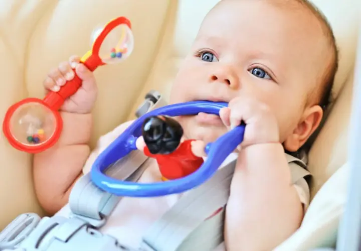 Bambino con sonaglio rosso in mano e gioco blu in bocca, esplora il mondo con i sensi. Ottimo per stimolare neonato ad afferrare oggetti.