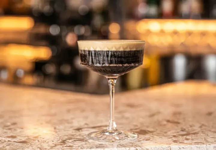 Un bicchiere di caffè martini con schiuma cremosa su un bancone di marmo, con luci sfocate sullo sfondo.