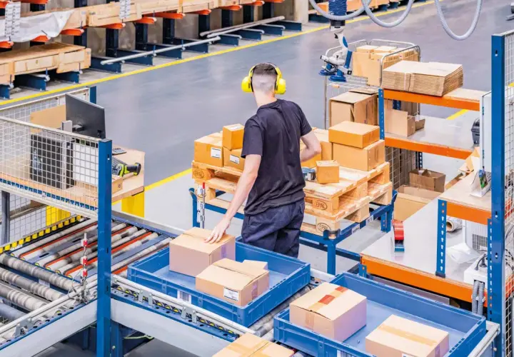 Ein Mitarbeiter mit Kopfhörern sortiert Pakete in einem Lager. Hier wird alles **assemble to order** für den Versand vorbereitet.