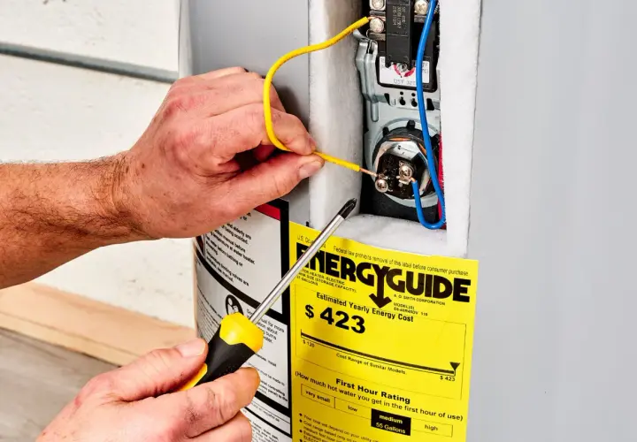 Ręce majsterkowicza podłączają przewody do bojlera, obok żółtej etykiety EnergyGuide, która informuje, ile prądu pobiera grzałka do bojlera.