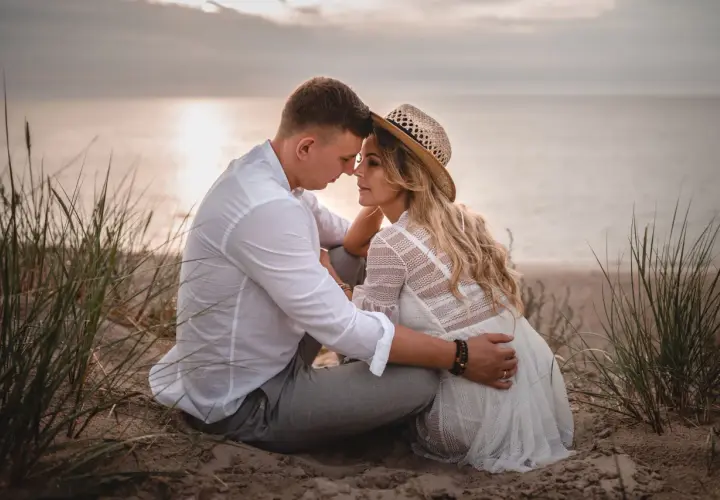 Romantyczna sesja we dwoje na plaży. Para siedzi blisko siebie, otoczona trawami, z widokiem na spokojne morze o zachodzie słońca.
