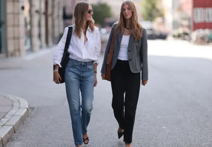 Dos mujeres caminan por la calle. Una, con un estilo demure, lleva vaqueros y camisa blanca. La otra, con chaqueta gris y pantalones negros, camina con elegancia.